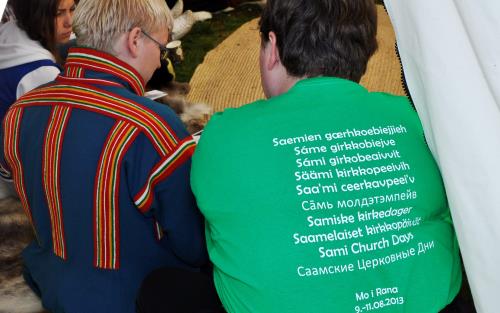 Två personer sitter bredvis varandra. Den ena har traditionell samisk dräkt. Den andra har en grön T-Shirt med texten "Samiska kyrkodagar" skrivet på et mängd olika språk.