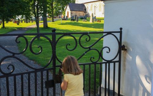 Flicka står vid svarta järngrindar. På andra sidan grinden fortsätter en grusgång upp till en stenkyrka.