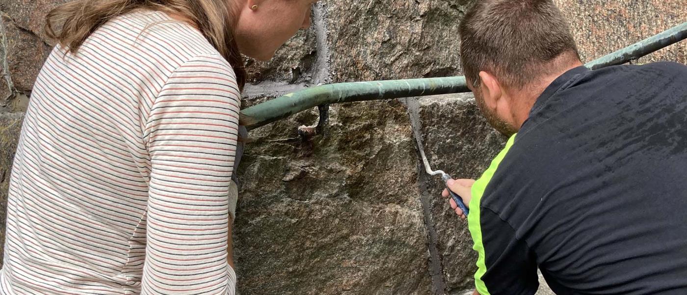 En kvinna och en man sitter på huk intill en mur av naturstenar. Mannen fyller i en fog med fogbruk mellan stenarna.