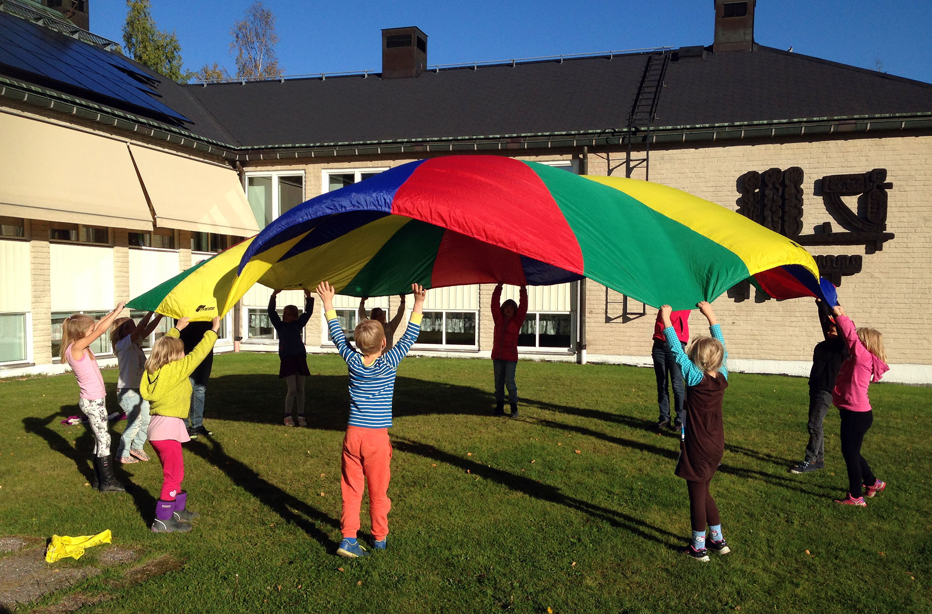 Tio barn och en ledare leker med en stor färggrann "fallskärm". De hjälps åt att kasta upp den i luften.De är utomhus på en gräsmatta.