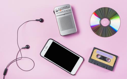 Olika musikmedier: en smartphone, ett kassettband, en cd-skiva och en fm-radio. Alla liggande på en rosa bakgrund.