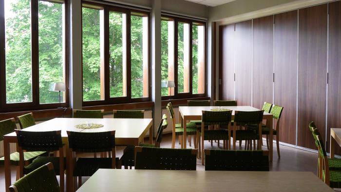 Rum med många fönster och flera kvadratiska bord med gröna stolar.