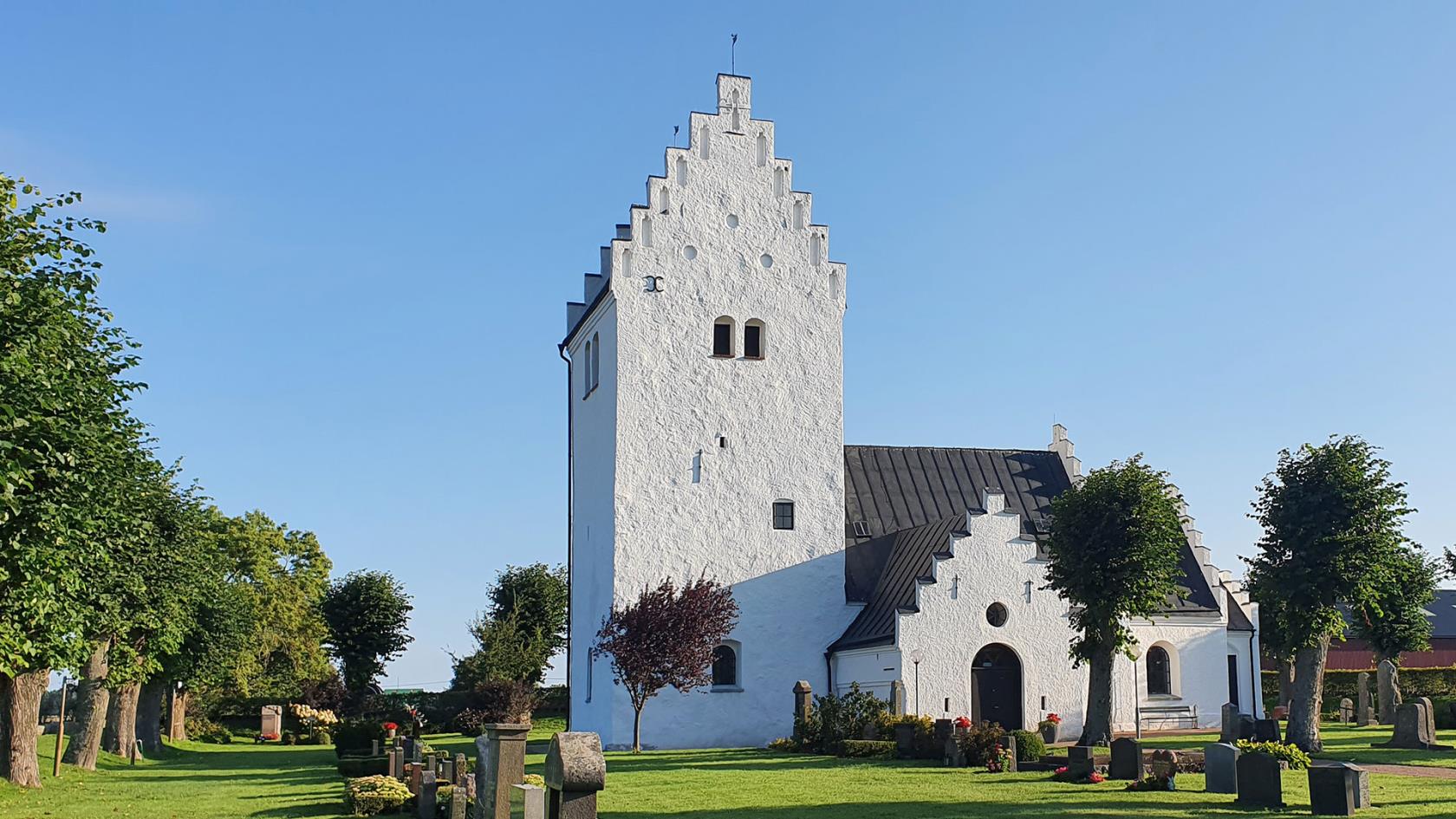 Vit kyrka med grönskande kyrkogård framför belyst av sensommarsol