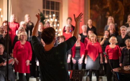 Körledare Malin Ekstrand syns bakifrån i skärpa, hon dirigerar kören. I oskärpa ser man en del av den stora kören Åby gospel. De är klädda i rött och svart.