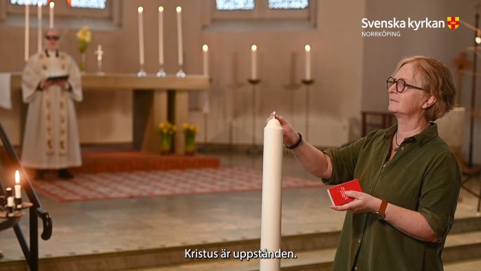 I förgrunden ser man musiker Mariette tända Kritusljuset. I bakgrunden ser man koret i Krokeks kyrka, smyckat med ljus, kors och påskliljor, och prästen Ida står vid sidan av altaret.