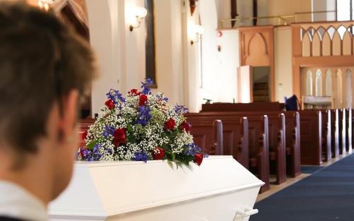 En vit kista med ett stort blomsterarrangemang står i kyrkan. En person i förgrunden kollar på kistan.