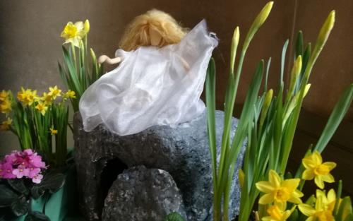 En docka med långt hår sitter på en sten och runt om är det påskliljor 