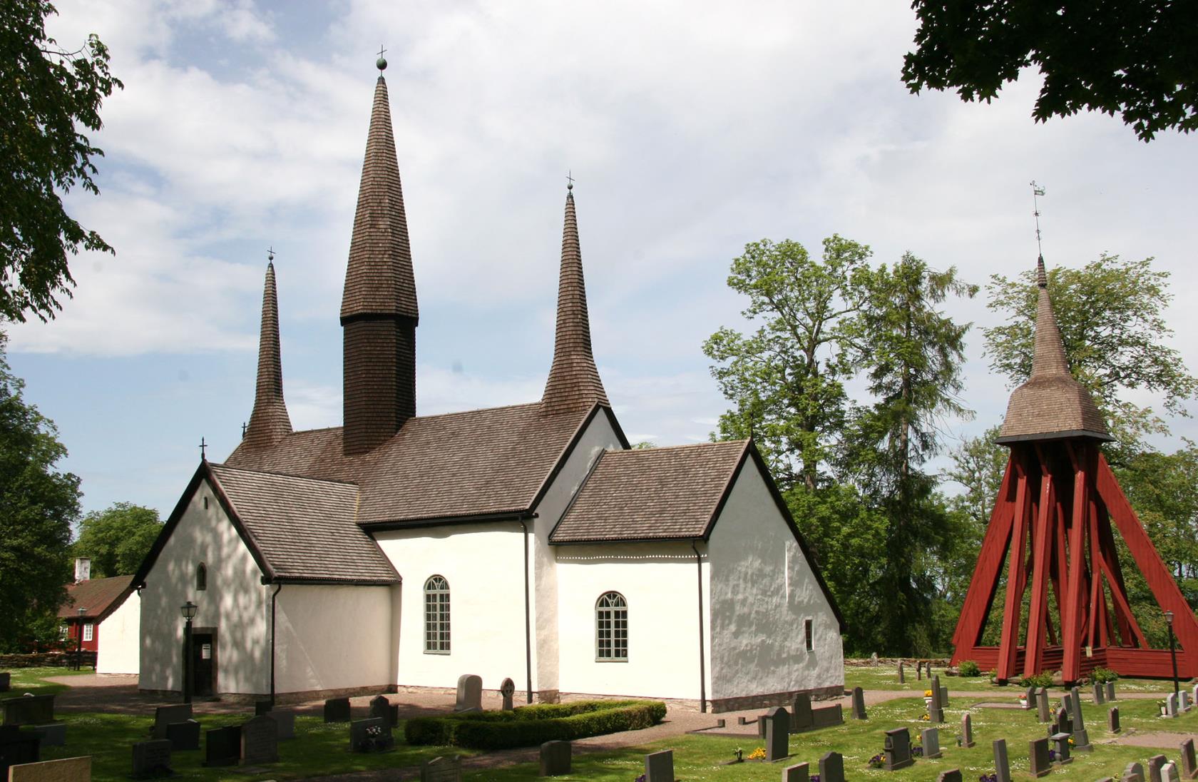 Kungslena kyrka med sina tre kyrktorn samt kyrkklockan intill, runt omkring är lummiga gröna träd och grönt gräs på kyrkogården.