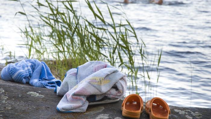 Ett par tofflor och två handdukar ligger på en klippa. Två personer badar i bakgrunden.