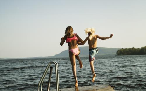 Två barn håller hand och hoppar från bryggan i vattnet.