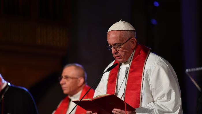 Påve Franciskus i vit dräkt och röd stola läser högt ur en bok i en mikrofon.