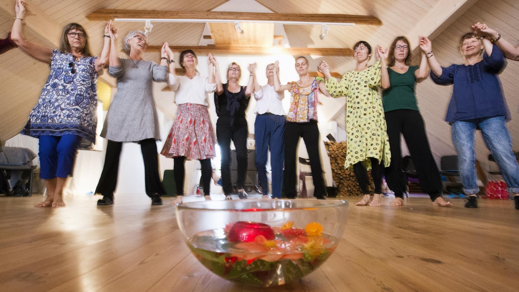 Några kvinnor dansar ringdans runt en skål med vatten och olika frukter som står på golvet.