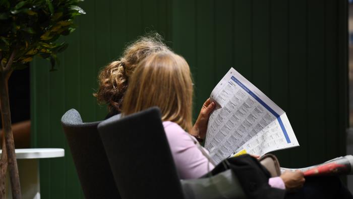 Två kvinnor sitter bredvid varandra inomhus och läser en tidning.