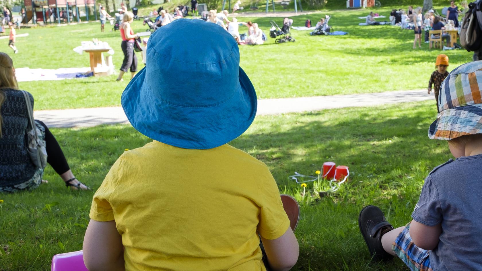 Två små barn med färgglada solhattar sitter i gräset i en park full av människor.