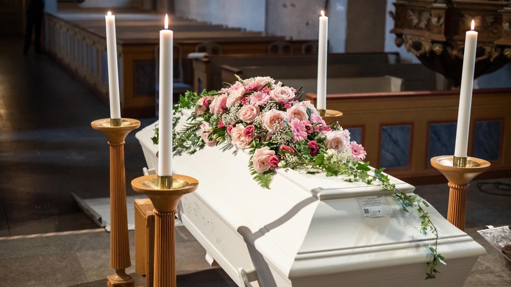 En vit kista med rosa blommor står omringad av ljus uppe vid koret.