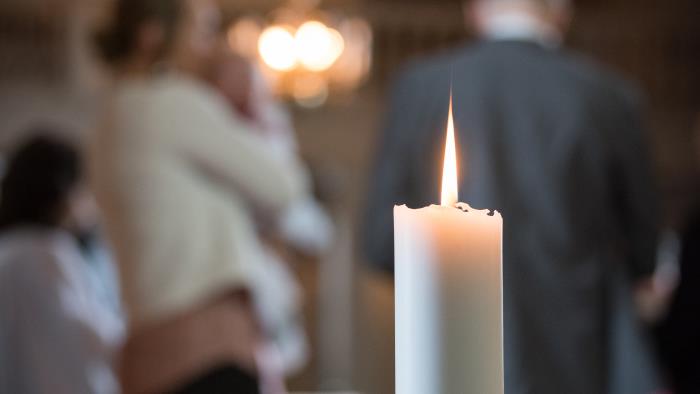 Ett ensamt ljus brinner i förgrunden. Suddiga personer i bakgrunden.