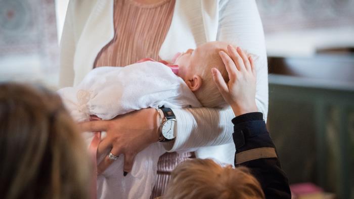 En bebis i dopklänning sover i någons famn. Ett barn klappar på bebisens huvud.