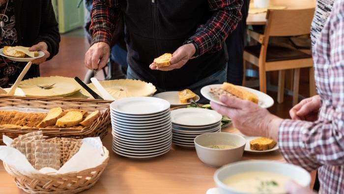 Runt ett fikabord står några personer och brer sina smörgåsar.