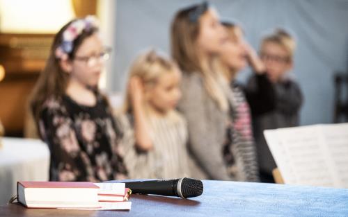 En psalmbok och en mikrofon ligger framlagda. I bakgrunden syns sjungande barn.