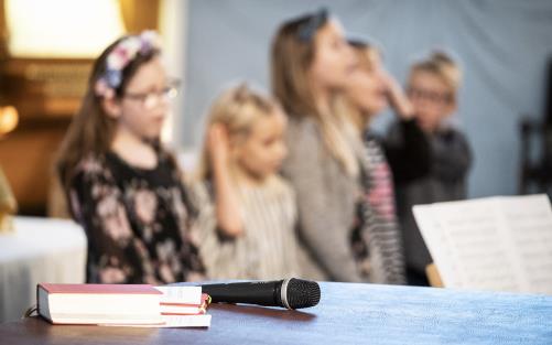 En psalmbok och en mikrofon ligger framlagda. I bakgrunden syns sjungande barn.