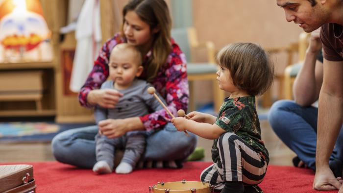 Ett litet barn spelar på en tamburin som ligger på en röd matta. Andra barn och vuxna sitter runt omkring.