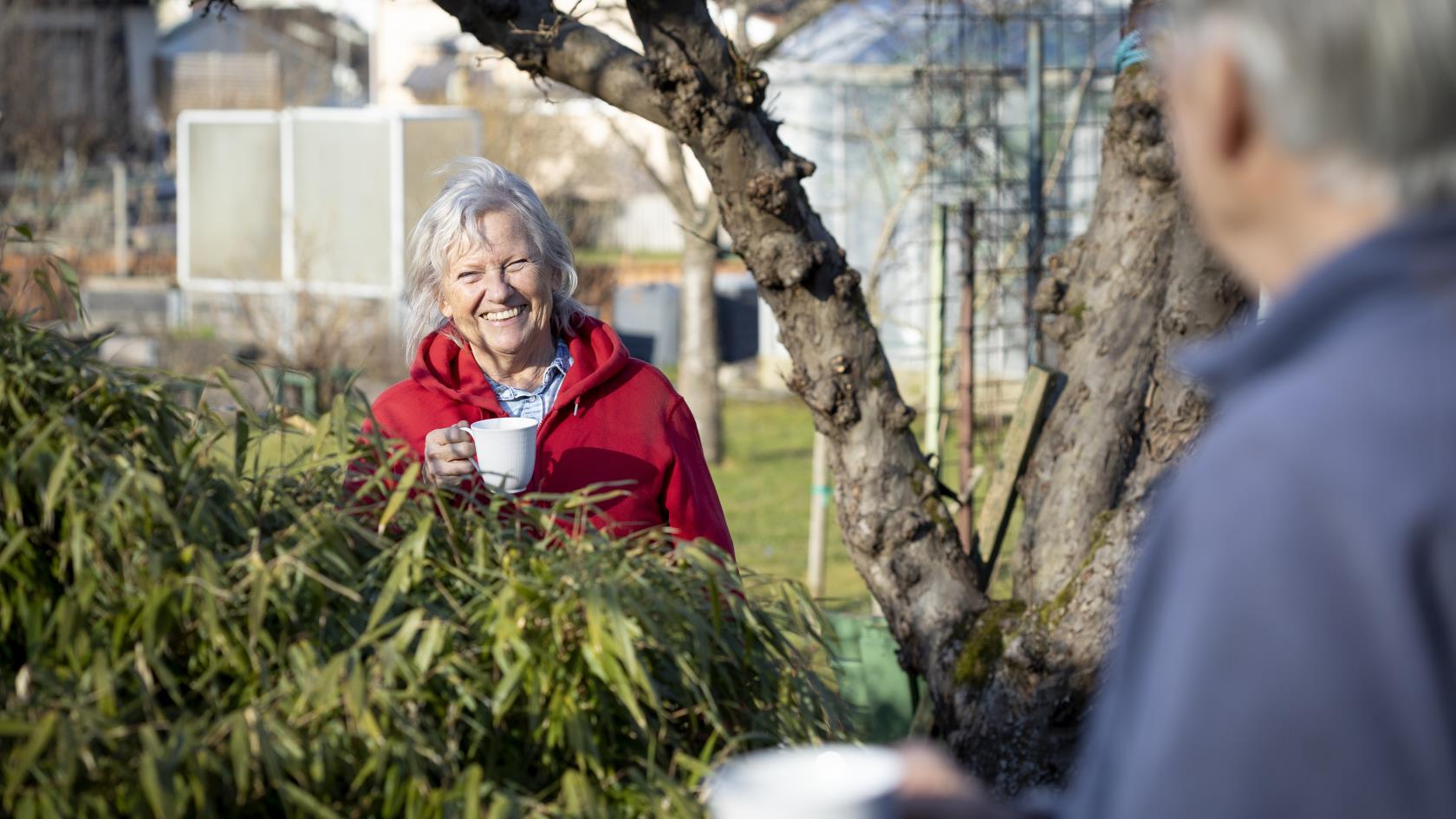 Leende kvinna med kaffekopp i handen står i sin trädgård och pratar med granne på andra sidan häcken.