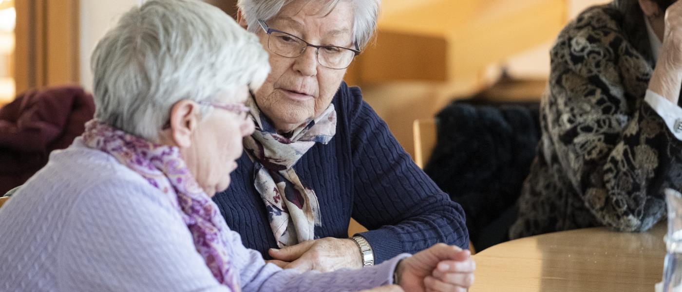 Två äldre kvinnor sitter bredvid varandra vid ett bord och pratar.
