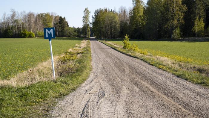 En grusväg mellan två åkrar. I vägkanten står en blå skylt med bokstaven M för mötesplats.