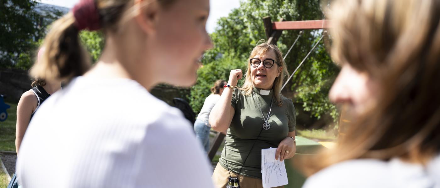 En kvinnlig diakon pratar med ungdomar utomhus en sommardag.