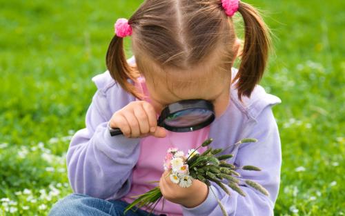 En liten flicka med förstoringsglas sitter på gräset och studerar buketten hon just plockat. 