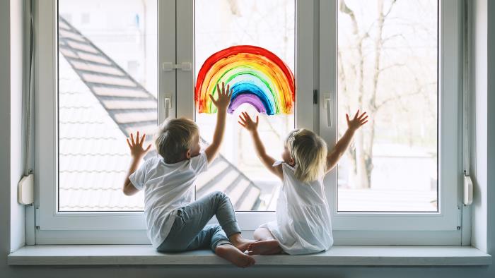 Två barn sitter vid ett fönster. På fönstret är en regnbåge målad.