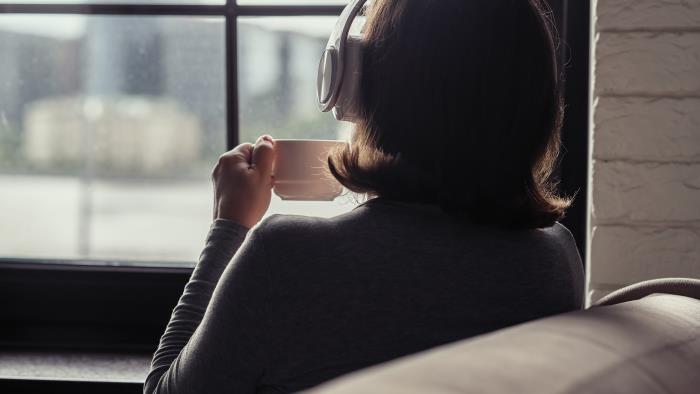 En kvinna med hörlurar sitter i en soffa med en kopp kaffe och tittar ut genom fönstret.
