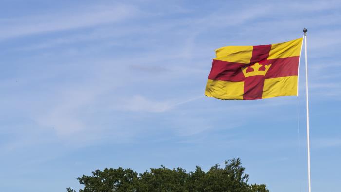 En flaggstång med Svenska kyrkans flagga mot blå himmel och några trädkronor.