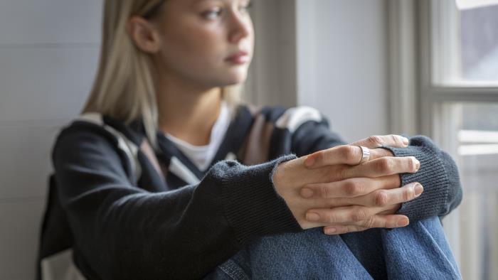 En tonårstjej sitter vid ett fönster med händerna om knäna.