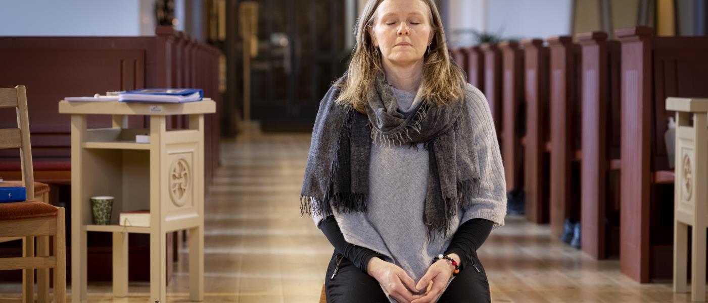 En kvinna sitter på en bönepall på en kyrkas golv, med knäppta händer och slutna ögon.
