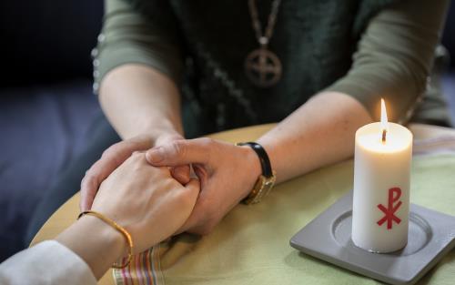 En diakon håller en annans hand i sina händer. Ett stearinljus med kristusmonogram brinner på bordet mellan dem.