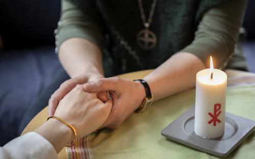 En diakon håller en annans hand i sina händer. Ett stearinljus med kristusmonogram brinner på bordet mellan dem.