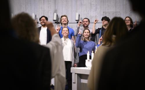 En blandad ungdomskör sjunger i en kyrka. De håller upp sina pekfingrar.