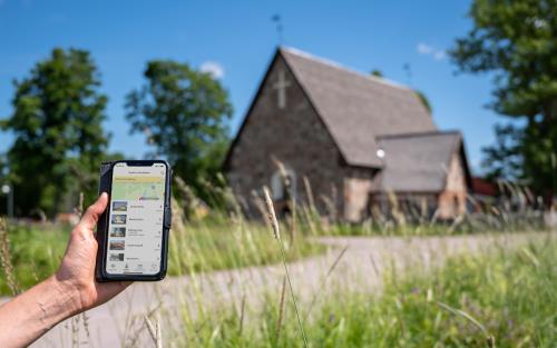 En person som kommit fram till en gammal kyrka håller en mobiltelefon i handen. På skärmen visas information om kyrkan.