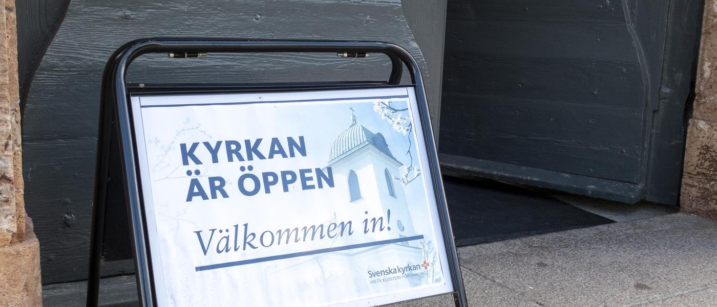 En skylt med texten Kyrkan är öppen välkommen in, står utanför en port.