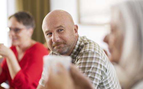 En man i rutig skjorta ler mot kvinnan bredvid. En kvinna med röd blus syns suddigt i bakgrunden.