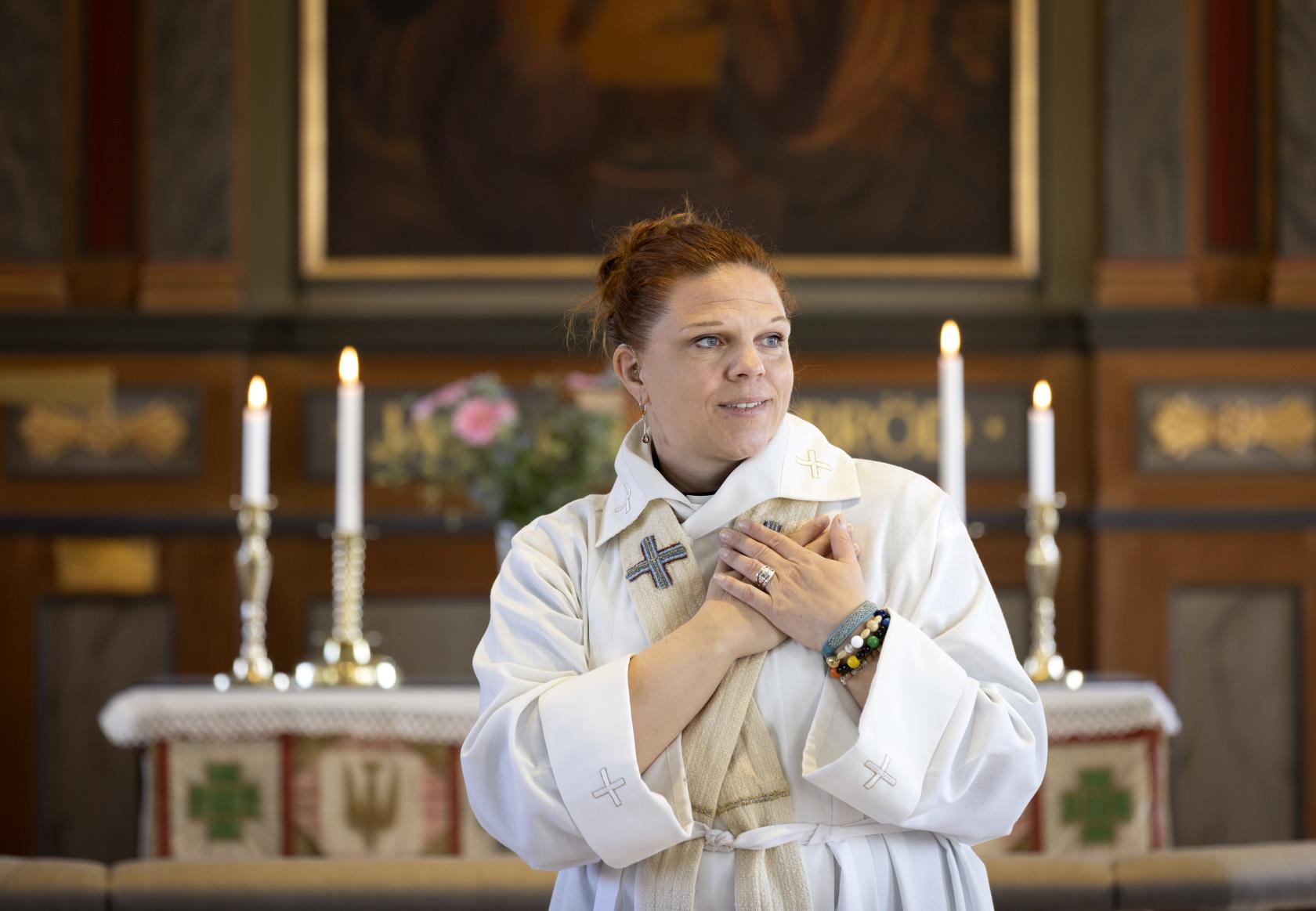 En kvinnlig präst i vit kåpa står vid koret och håller båda händerna mot hjärtat.
