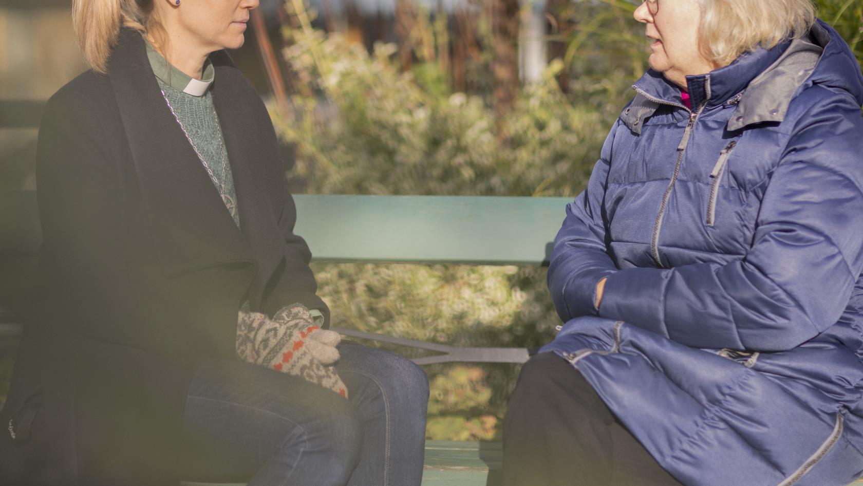 En kvinnlig diakon och en äldre kvinna sitter utomhus på en bänk och samtalar.