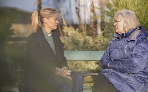 En kvinnlig diakon och en äldre kvinna sitter utomhus på en bänk och samtalar.