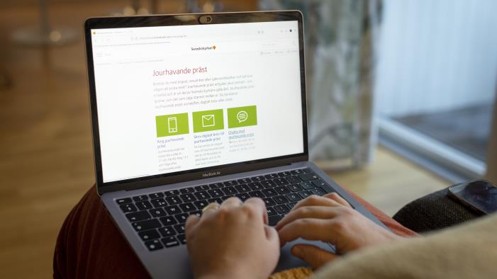 En person sitter vid med en bärbar dator i knät och läser på Svenska kyrkans webbsida om jourhavade präster.