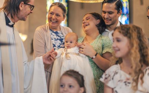 Ett dop i en kyrka. En kvinna med sin man bakom sig, håller barnet i famnen och skrattar. En manlig präst håller barnets hand.