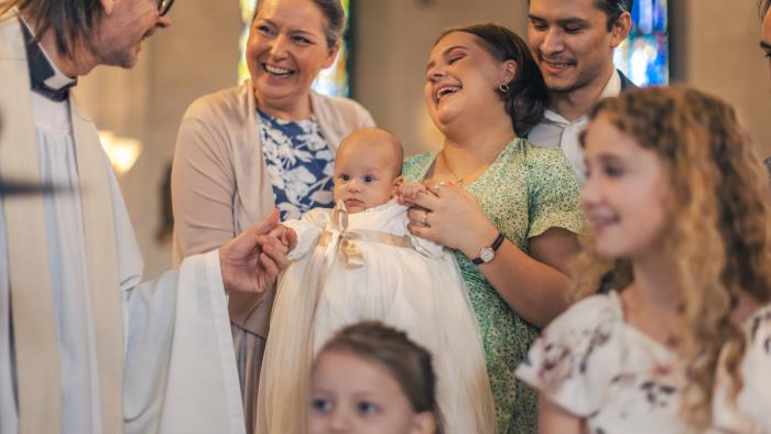 Ett dop i en kyrka. En kvinna med sin man bakom sig, håller barnet i famnen och skrattar. En manlig präst håller barnets hand.