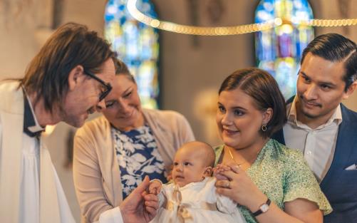 Ett dop i en kyrka. En kvinna med sin man bakom sig, håller barnet i famnen. En manlig präst håller barnets hand.