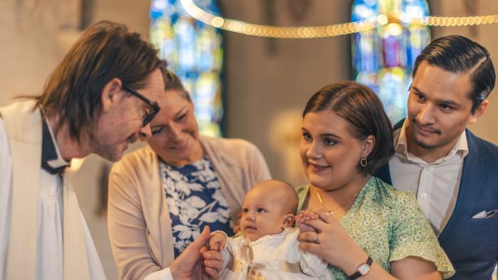 Ett dop i en kyrka. En kvinna med sin man bakom sig, håller barnet i famnen. En manlig präst håller barnets hand.