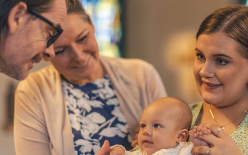 Ett dop i en kyrka. En kvinna med sin man bakom sig, håller barnet i famnen. En manlig präst håller barnets hand.